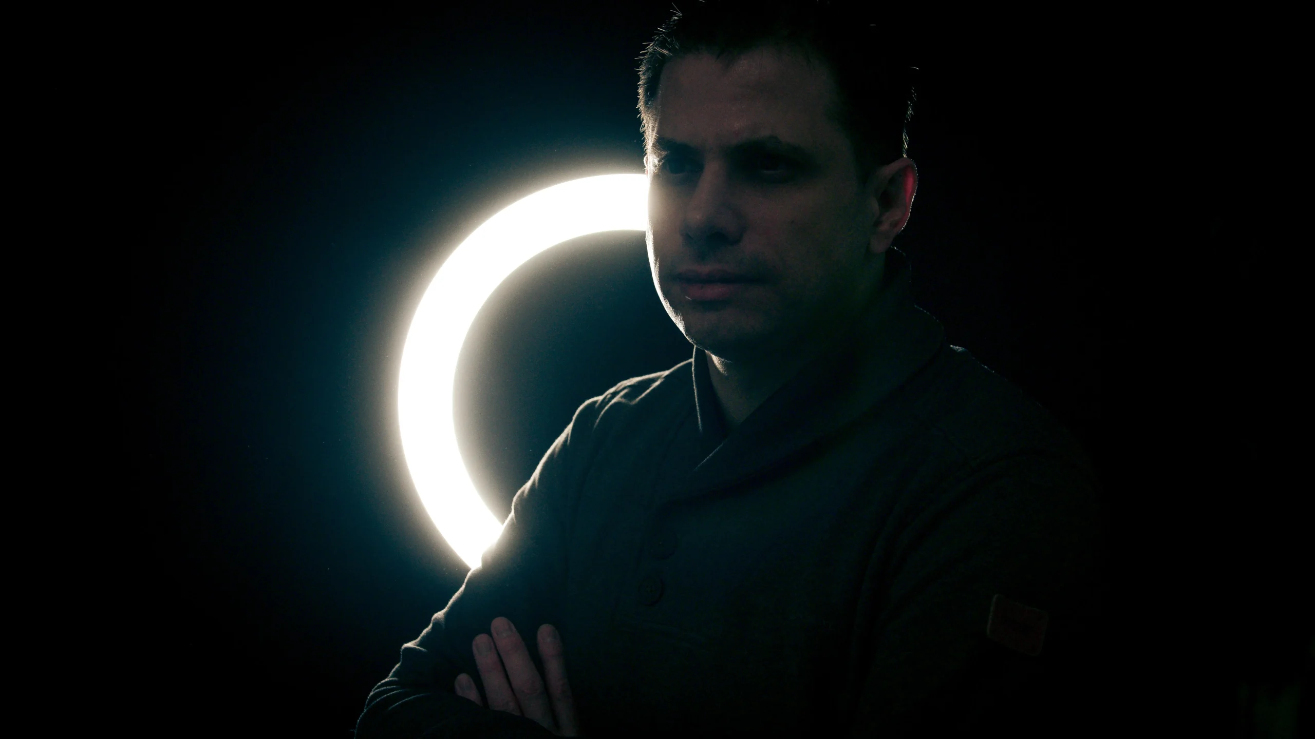 A male model in a darkened photo studio having his arms crossed for a standing pose with an illuminated ring light behind him.